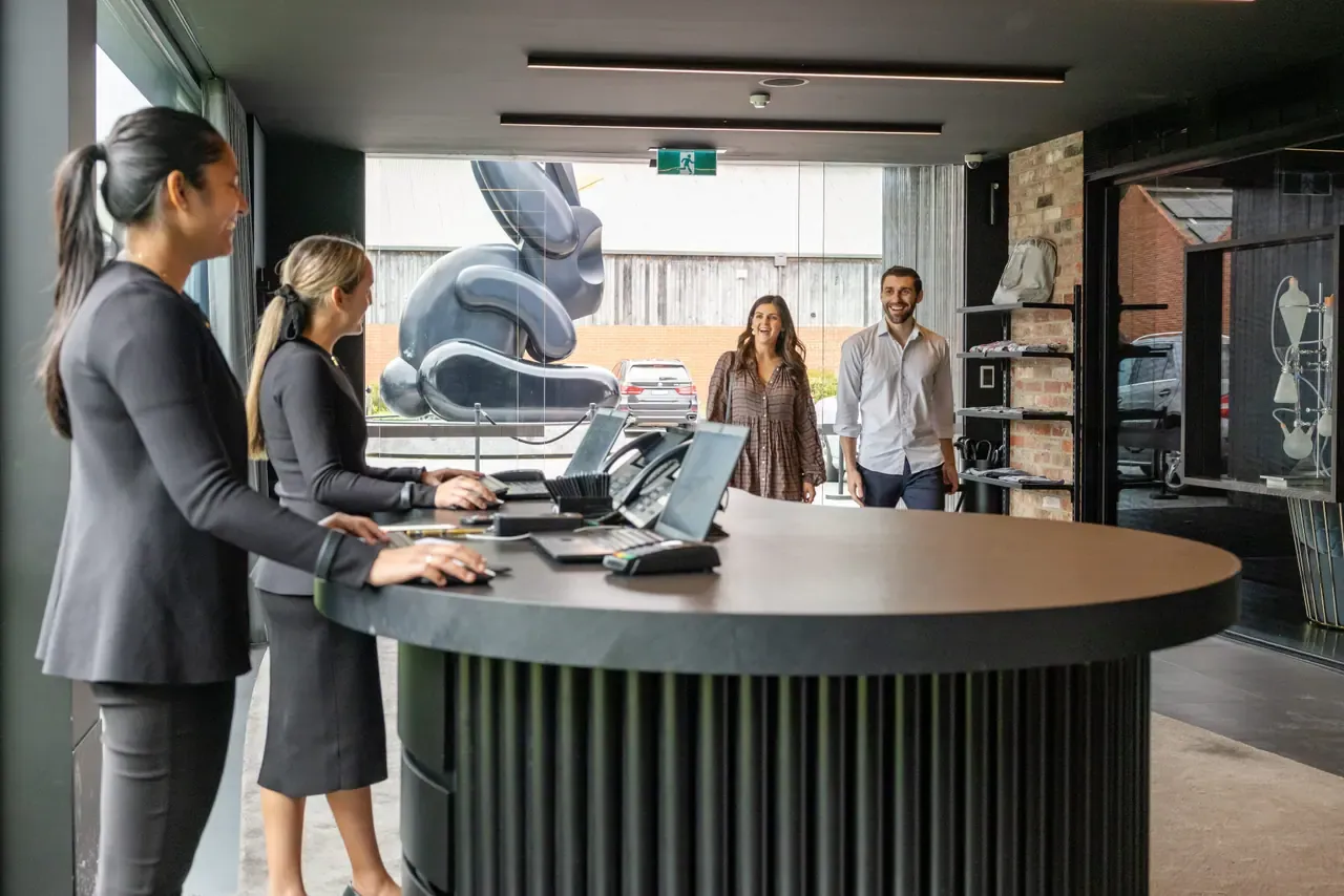 Staff at a curved reception desk greet two arriving guests at Jackalope Hotel, with the large dark jackalope sculpture outside