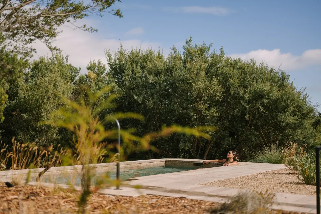 A bather resting at the end of a long stone-edged pool behind soft grasses and coastal scrub at Alba Thermal Springs