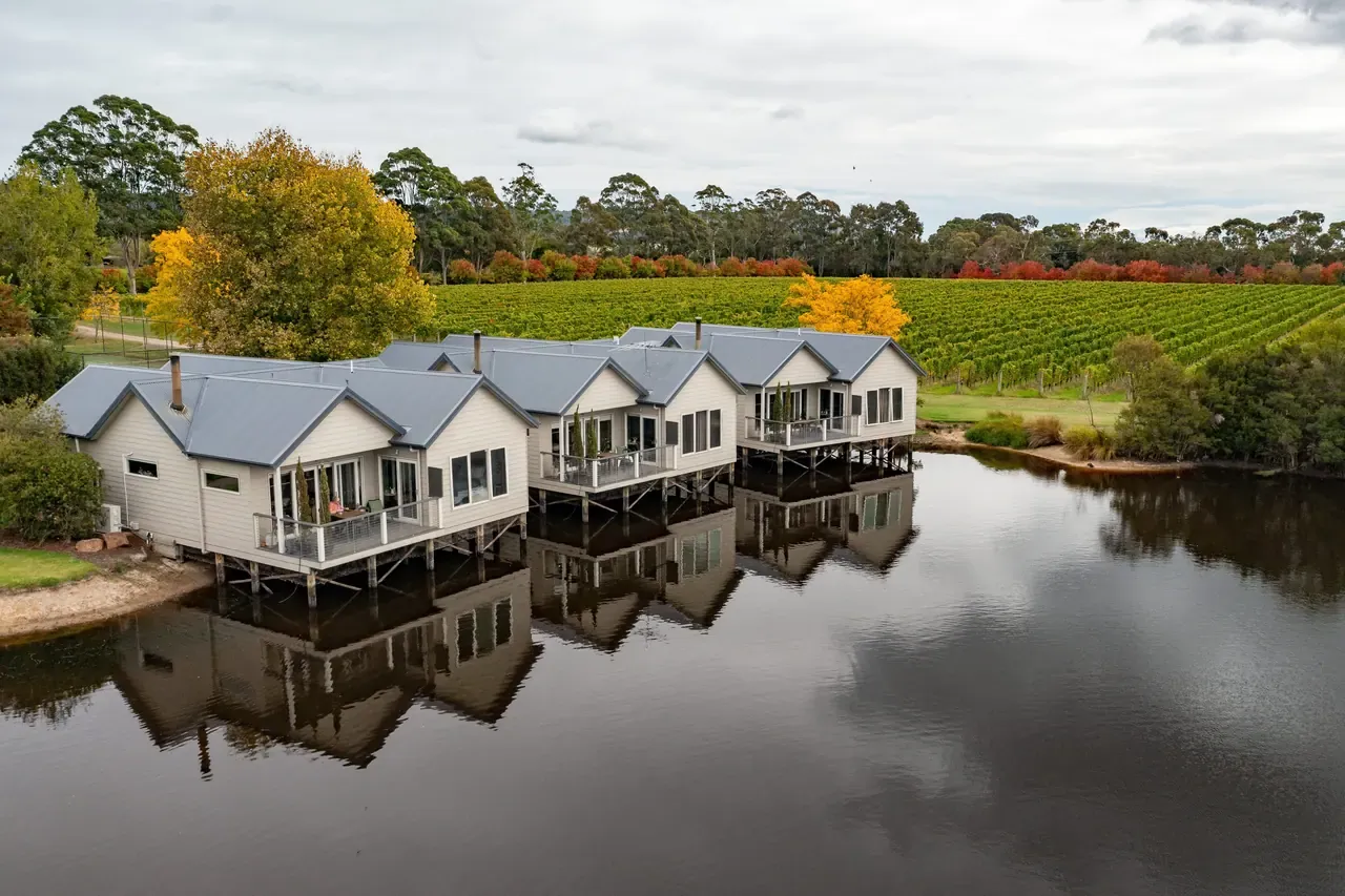 Row of grey-roofed cabins on stilts at Crittenden Estate reflected in a still lake, with vineyard and autumn trees behind