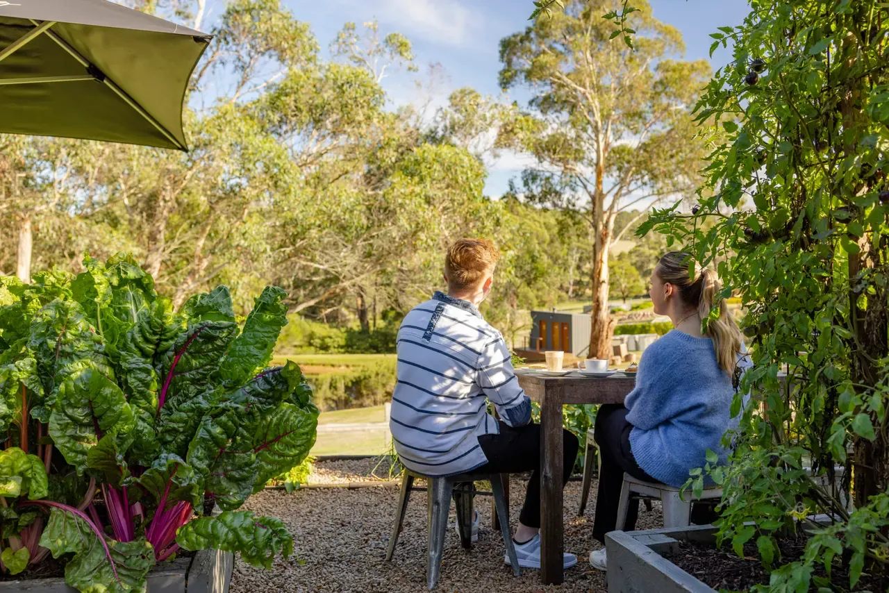 Two people seated at a garden table among vegetable beds, looking out over bushland at Green Olive at Red Hill