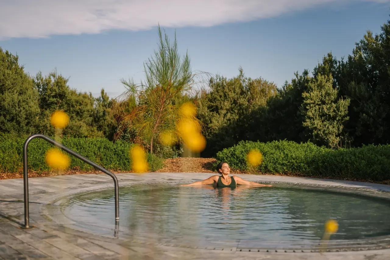A bather resting against the edge of a round thermal pool surrounded by coastal trees at Alba Thermal Springs