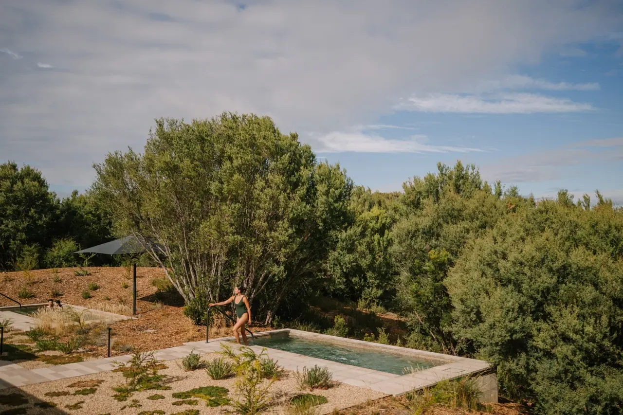 A bather climbing into a rectangular plunge pool tucked among tea-tree scrub at Alba Thermal Springs