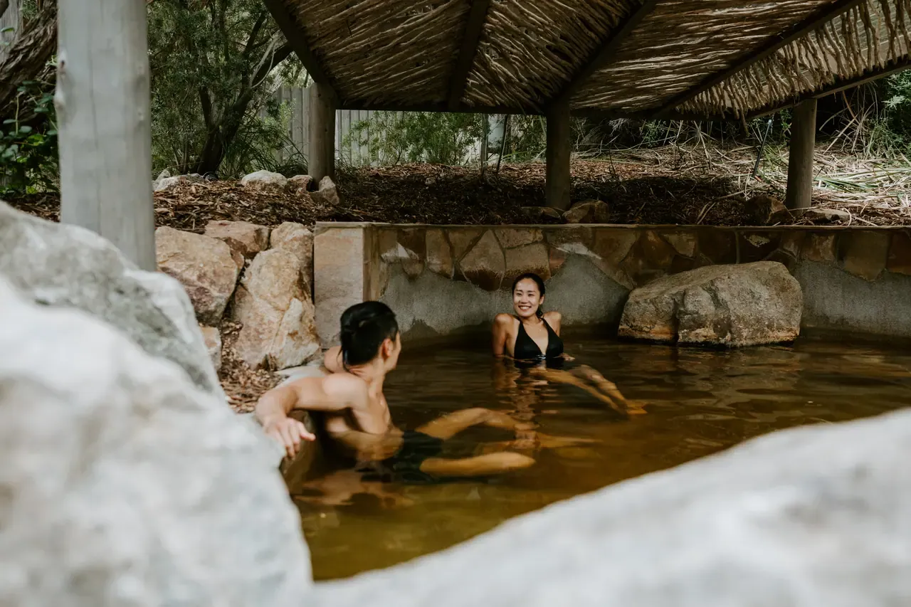 Two bathers chat in an amber-coloured thermal pool beneath a thatched shelter, framed by pale boulders at Peninsula Hot Springs