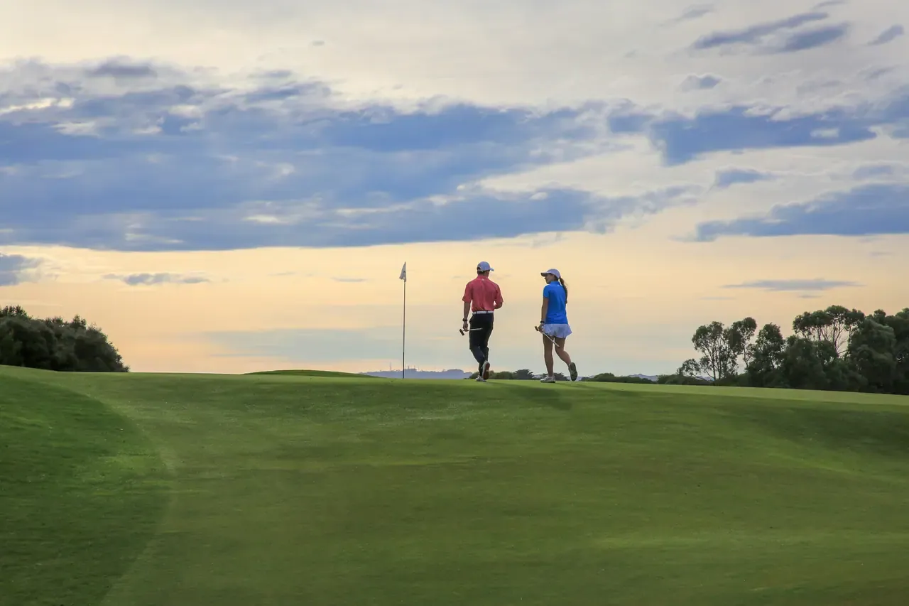 Two golfers walk over a grassy rise towards a flag against a soft pink and blue sunset sky.