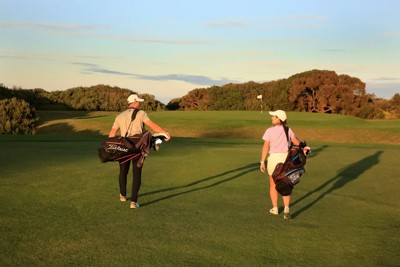 Two golfers walk with their bags up a fairway at Flinders Golf Course towards a flag, casting long shadows in evening light