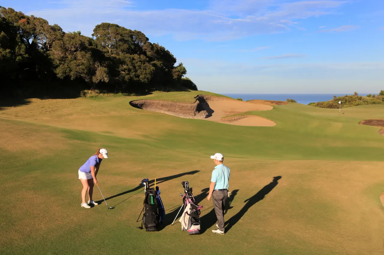 A golfer chips toward the green at The National Golf Club as a partner watches, with a large bunker and the sea beyond