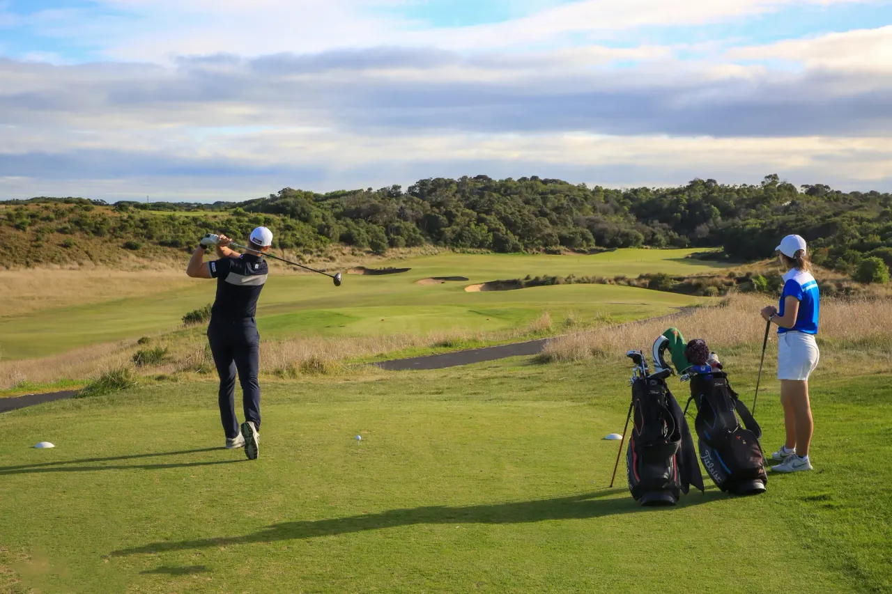 A golfer tees off at St Andrews Beach Golf Course as a second player waits beside two golf bags, with dunes and scrub behind