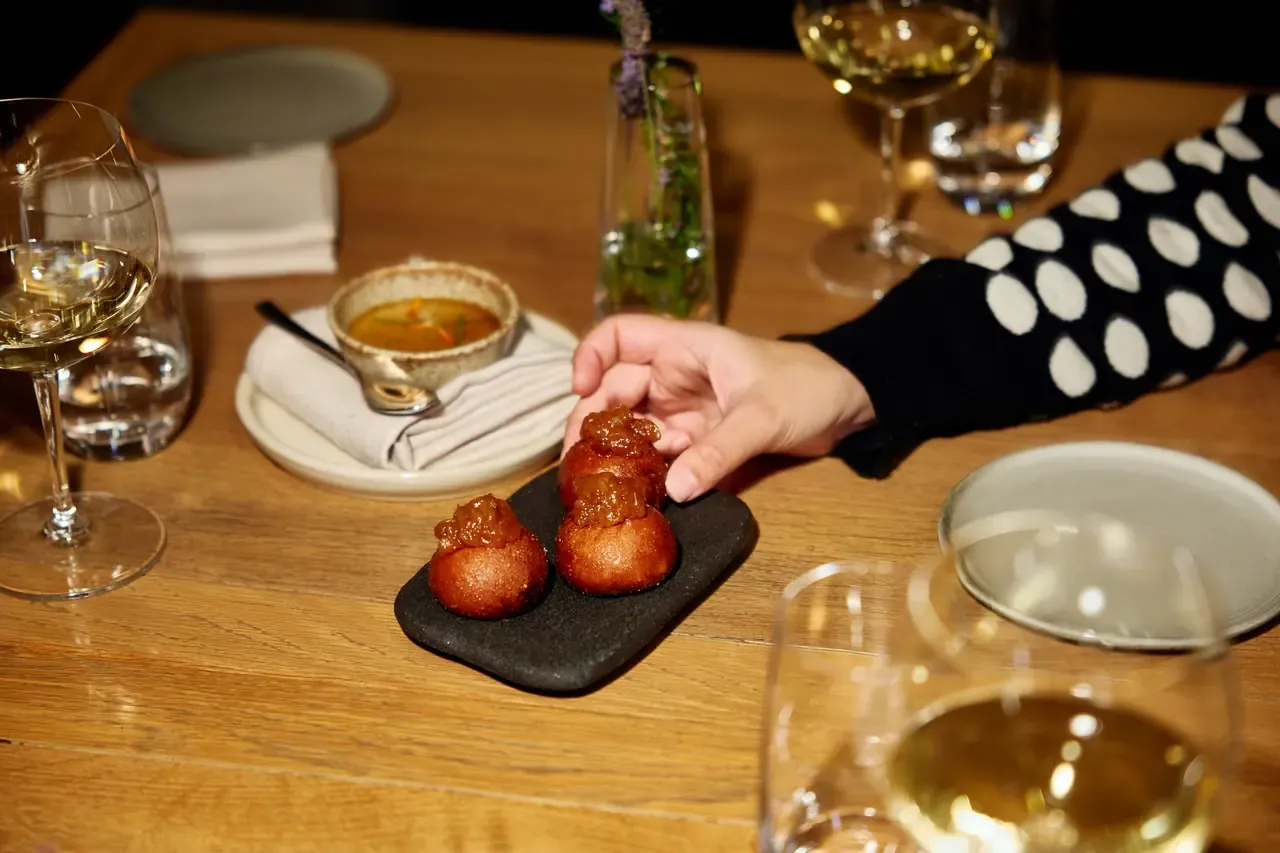 A diner picks up a glazed round snack from a slate board beside a small bowl of broth and white wine at Lindenderry