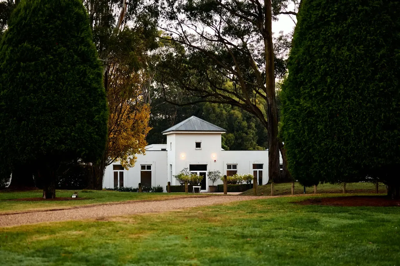 The white homestead at Lindenderry at Red Hill framed by tall cypress hedges, with a gravel drive and green lawn at dusk