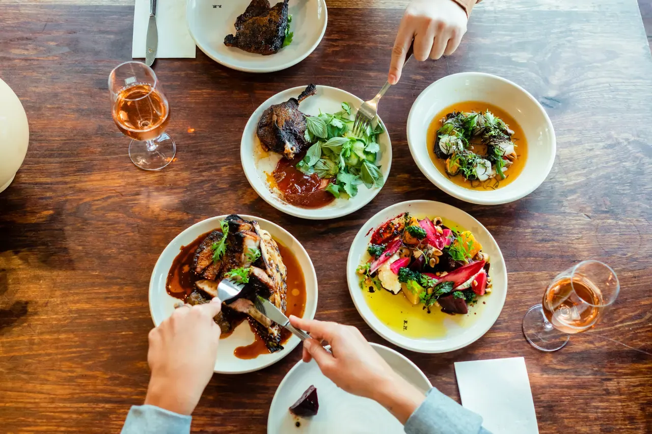 Overhead view of diners sharing roast duck, grilled meat, a beetroot salad and rose at a timber table