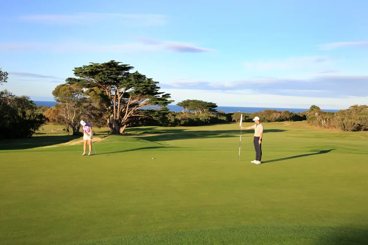 A golfer chips onto the green at Flinders Golf Course while another holds the flag, with cypress trees and ocean behind