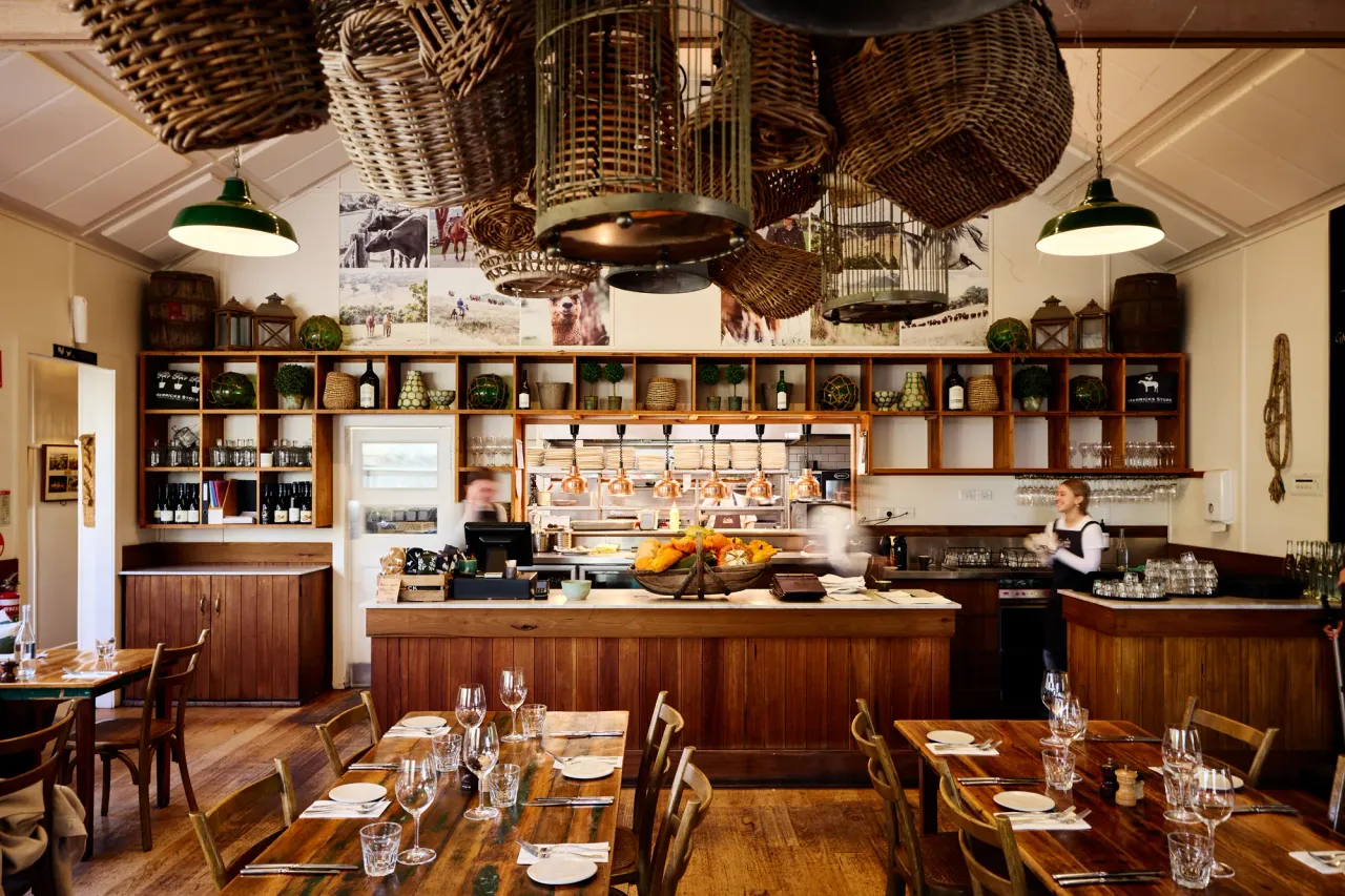 Timber tables set for lunch before an open kitchen with hanging woven baskets and shelves of wine at Merricks General Wine Store