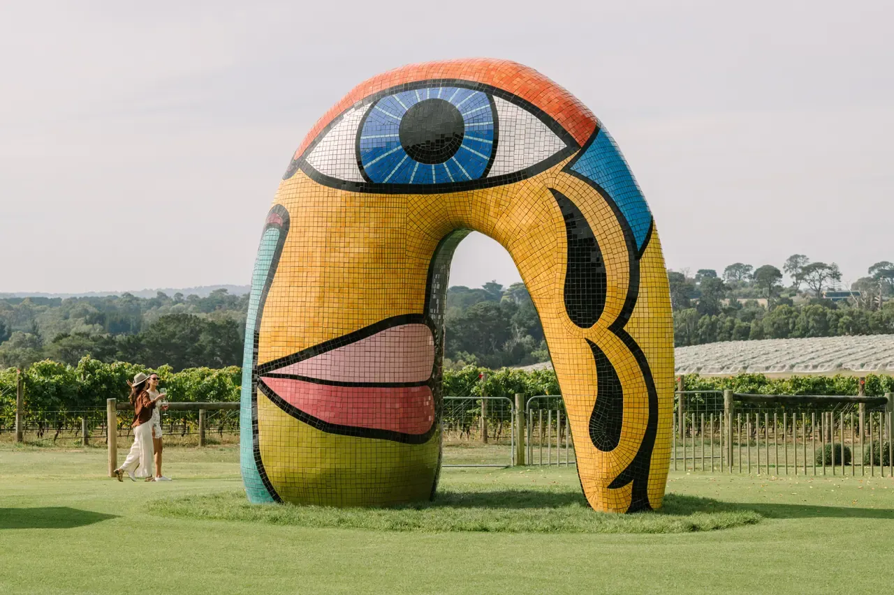 Two visitors walk past a large mosaic sculpture of a face with a single blue eye on the lawn at Pt. Leo Estate