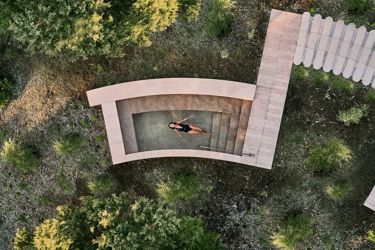 Overhead view of a bather floating in a curved stone plunge pool set among native shrubs, reached by a pale timber walkway