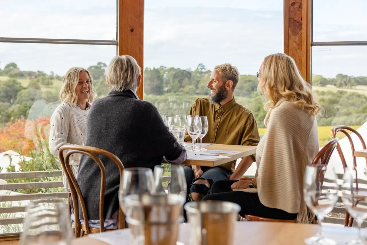 Four friends talking over wine glasses at a table by tall windows looking out to green hills at Montalto