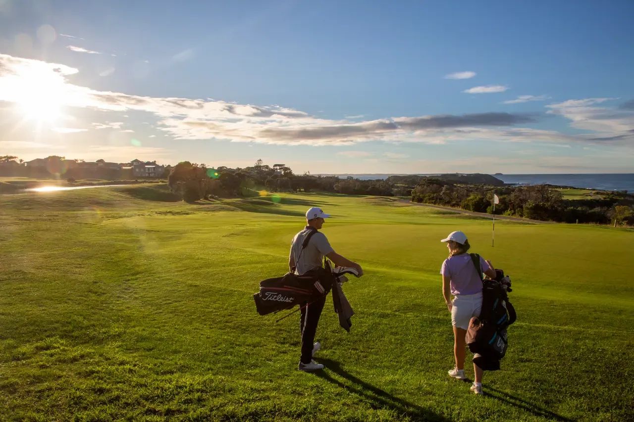 Two golfers carrying bags across a fairway at Flinders Golf Course towards the sea, with low sun flaring over the clubhouse