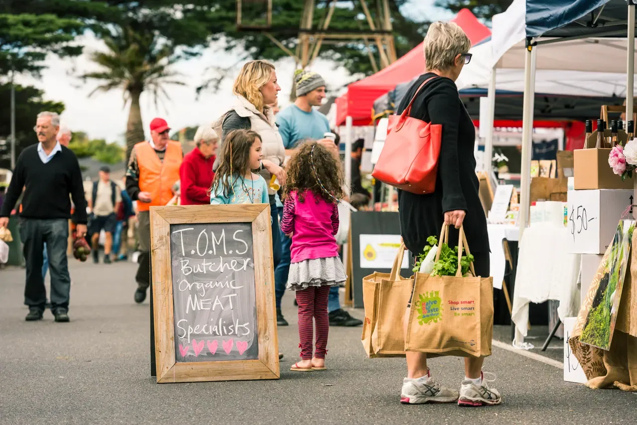 Families with shopping bags browse the marquees at Mornington Farmers Market, with Norfolk pines behind