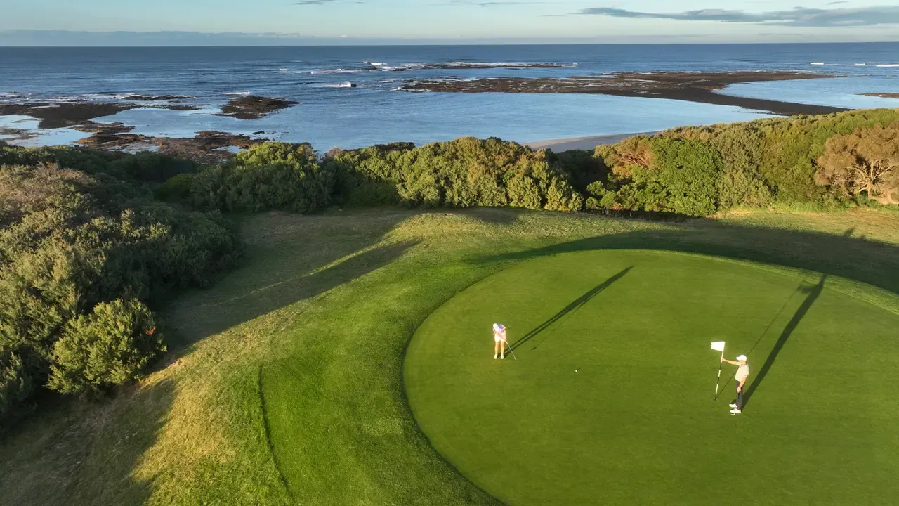 Two golfers on a putting green, one holding the flagstick, with coastal scrub and a rocky shoreline beyond in low golden light.