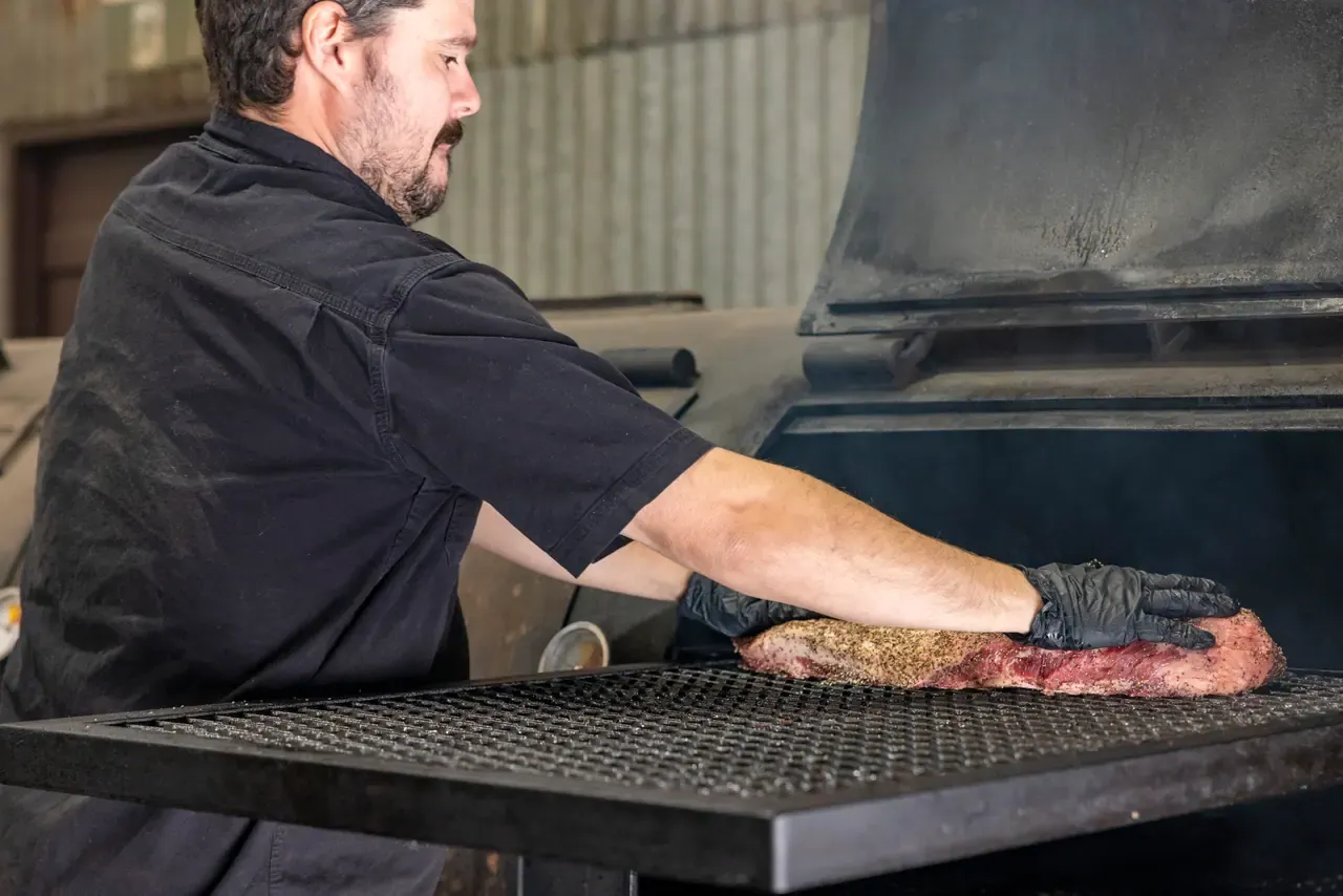A pitmaster in black gloves lays a seasoned brisket onto the grate of a large steel smoker