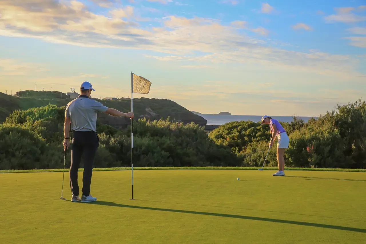 A golfer holds the flag while a second player putts on a green at Flinders Golf Course, with coastal scrub and a headland behind.