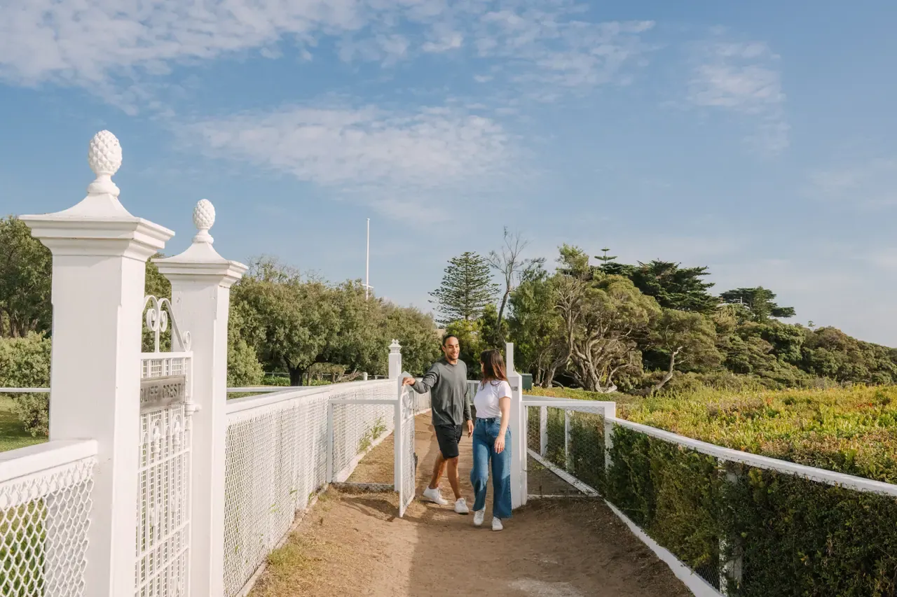 A couple pause by an ornate white gate on the fenced path of Millionaire's Walk, with coastal trees and a clear sky behind