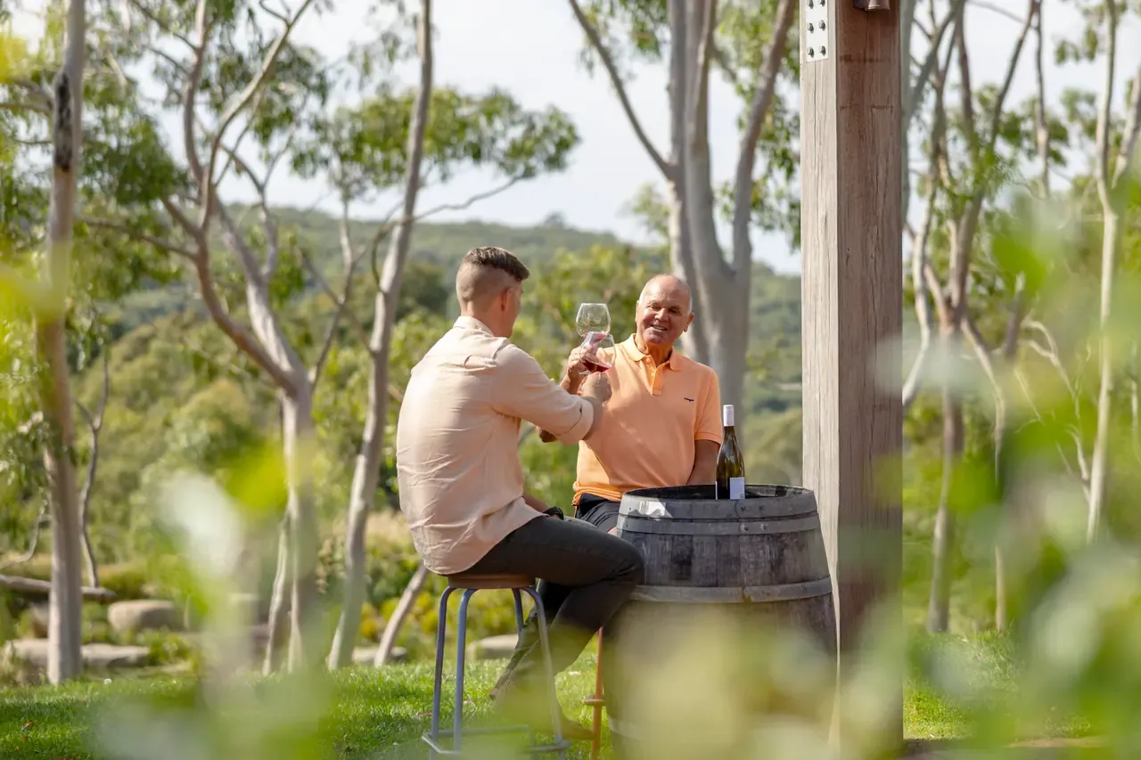 Two people sit on stools at a barrel table raising glasses of wine among gum trees and green hills