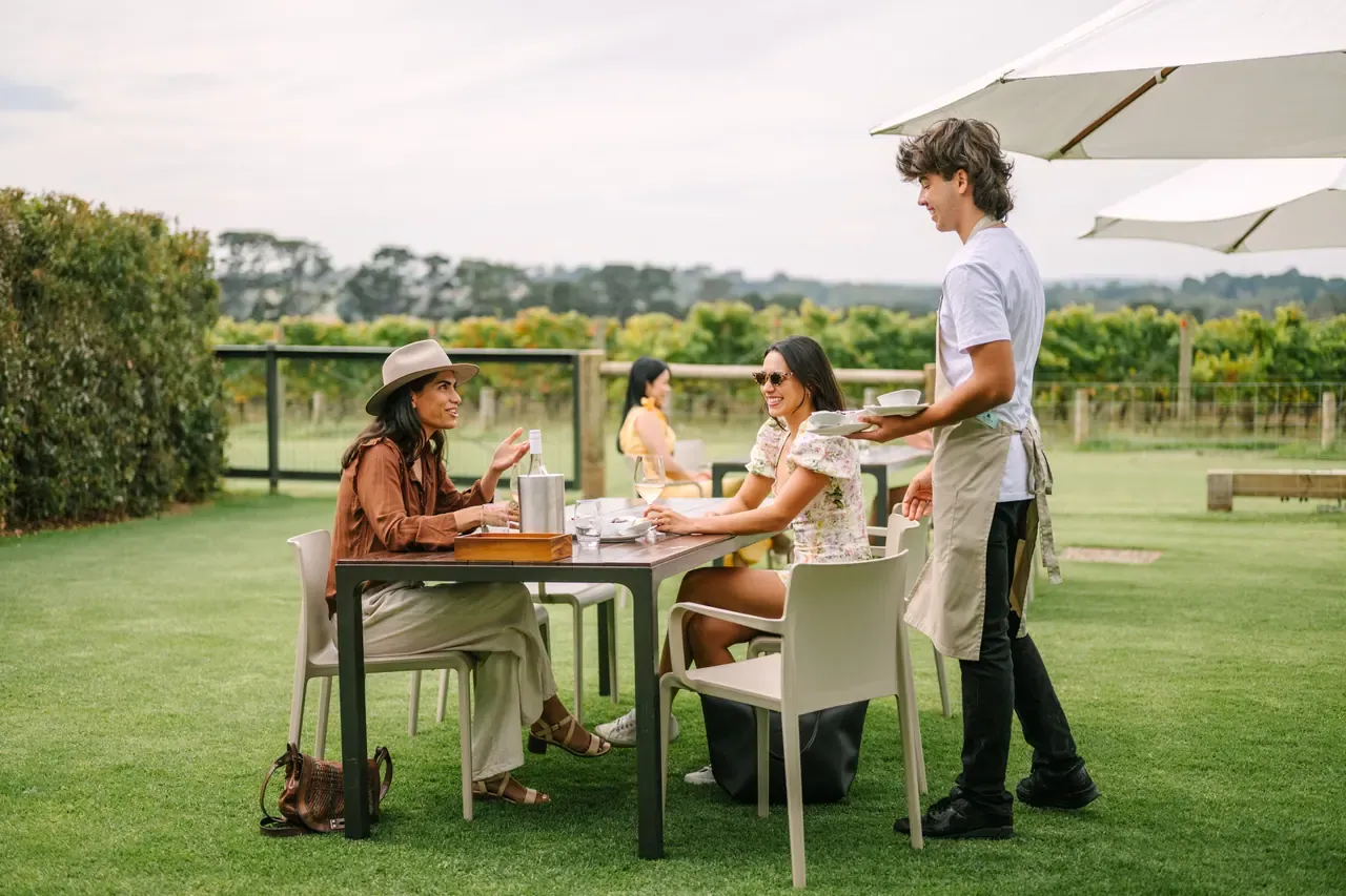 A server brings plates to guests at an outdoor table on the lawn at Pt. Leo Estate, with vineyard rows behind