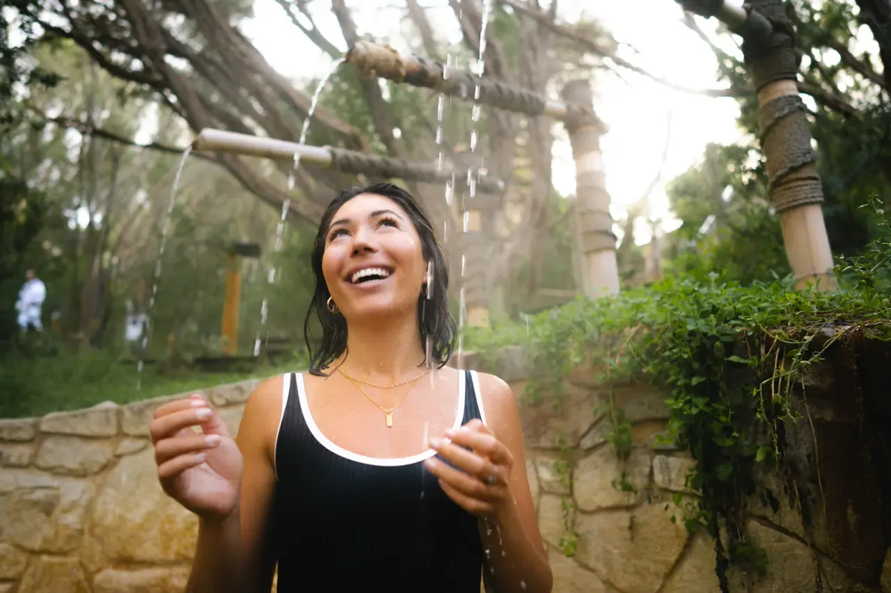 A smiling bather stands beneath streams of water from bamboo spouts at a rope-bound shower in the bush at Peninsula Hot Springs