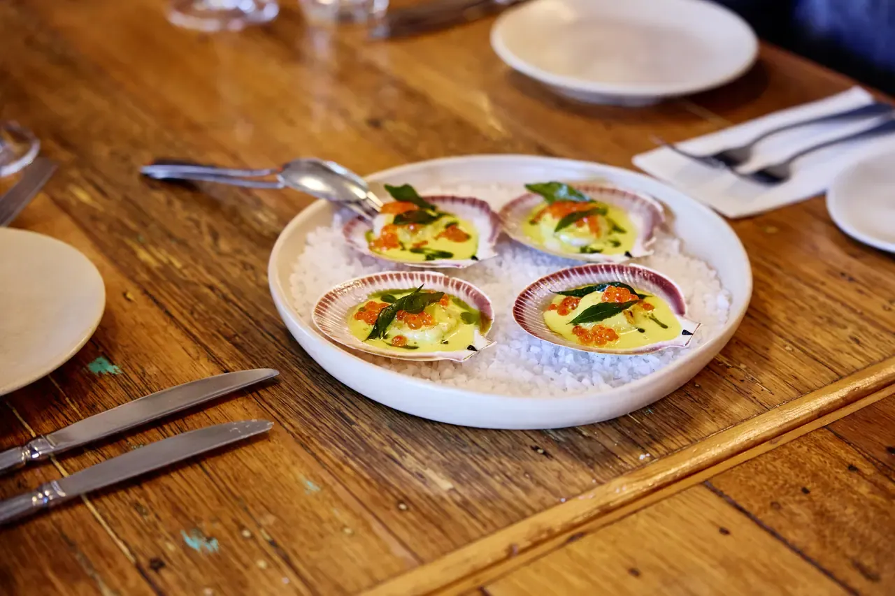 Four scallops on the half shell with a yellow sauce, orange roe and herbs, served on salt on a timber table