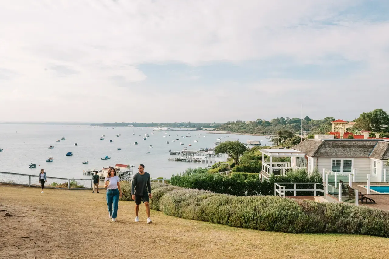 A couple walk across the clifftop lawn on Millionaire's Walk, other walkers behind, above a bay full of moored boats
