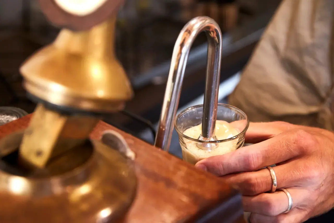 A small glass fills with frothy beer from a curved chrome spout beside a brass and timber beer engine