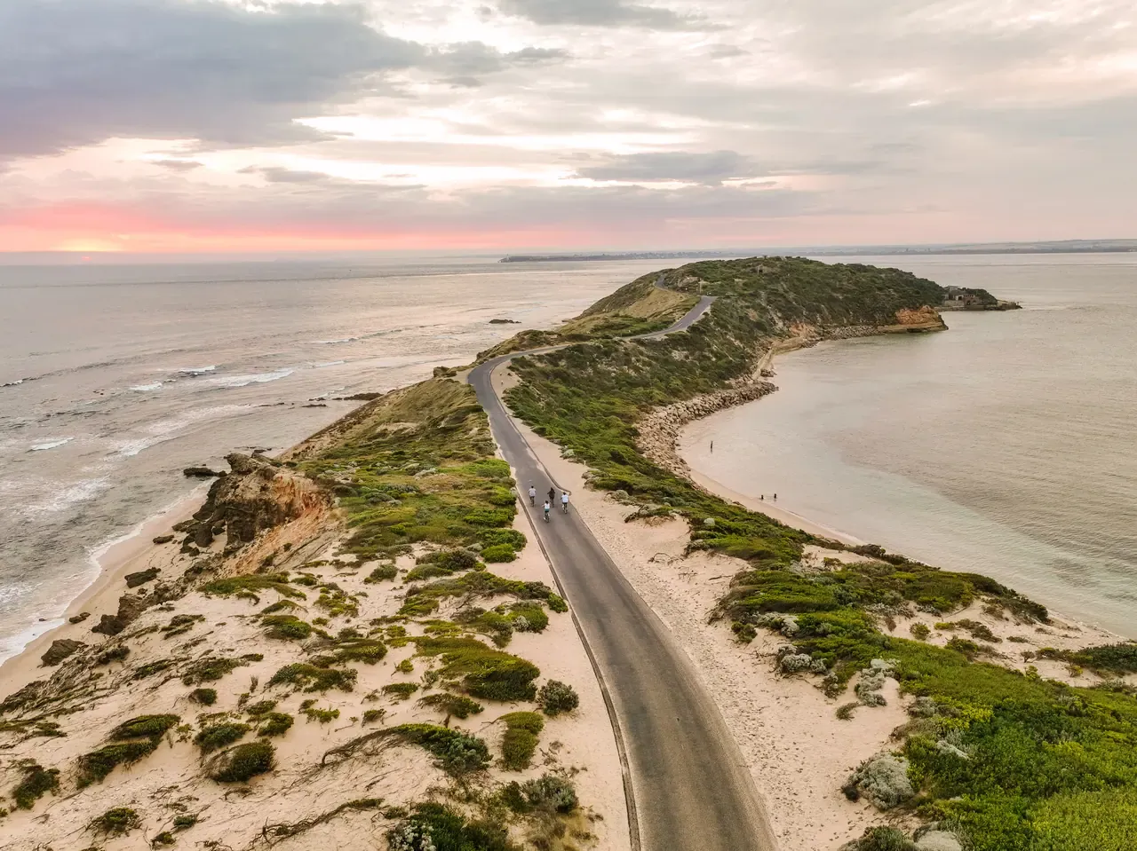 Aerial view of cyclists on the road running out along the narrow Point Nepean headland, with calm bay water and a pink sunset sky