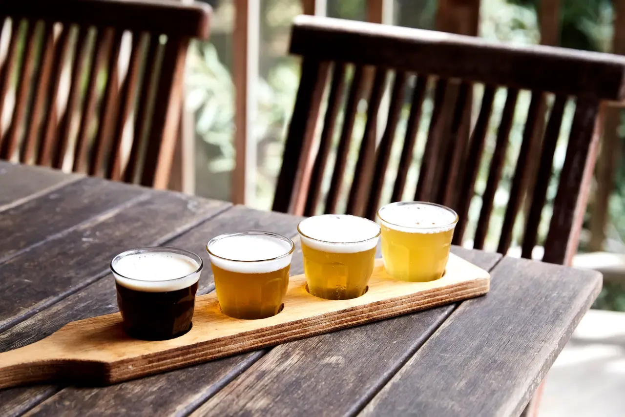 A tasting paddle of four beers, from dark to pale, on a weathered outdoor table beside timber chairs