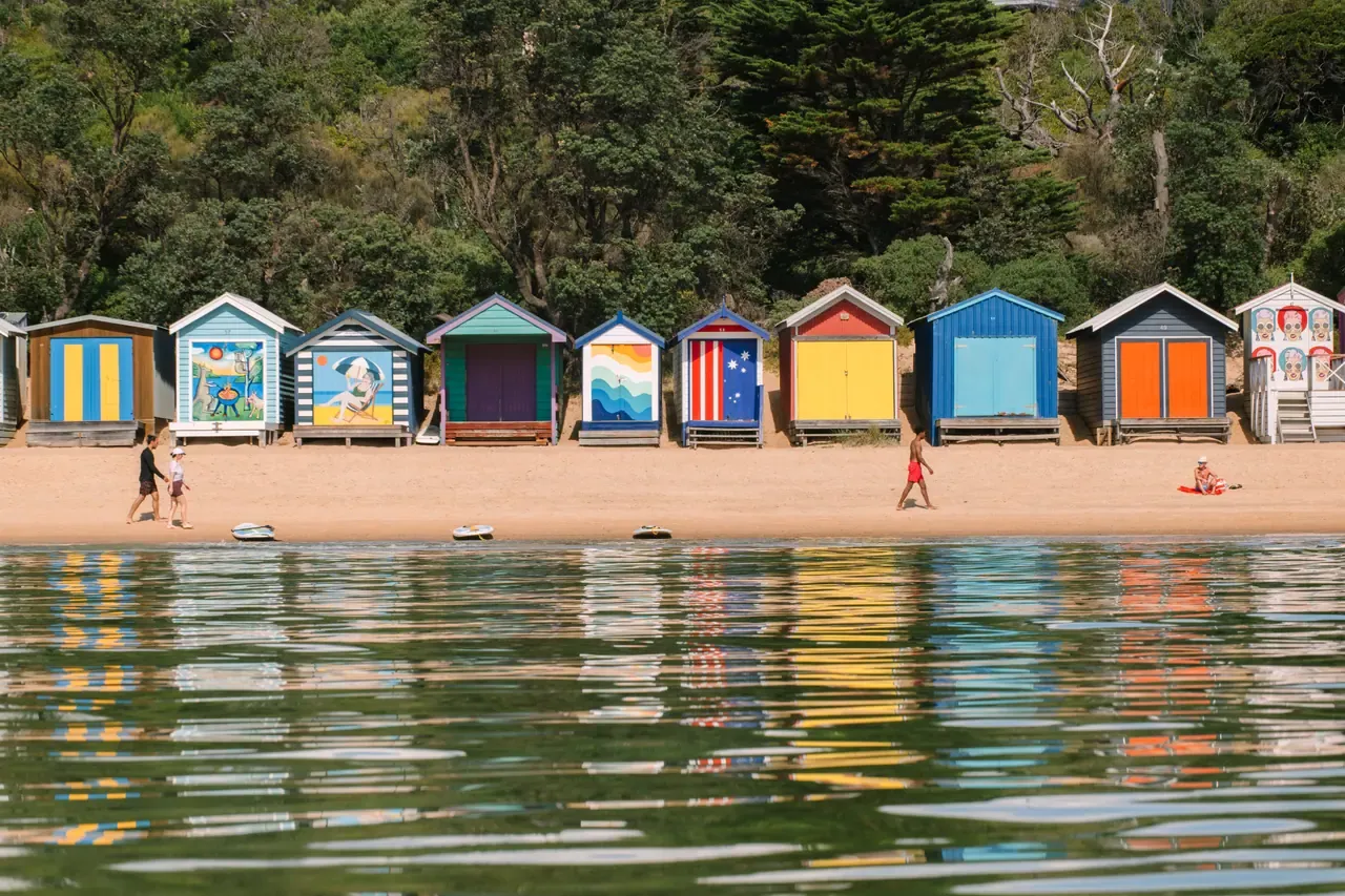 A row of colourful painted bathing boxes along the beach at Mornington, reflected in calm water, with a few beachgoers