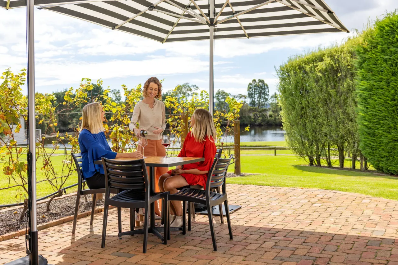 A host pours red wine for two guests at an umbrella-shaded table on a brick terrace overlooking vines and a lake