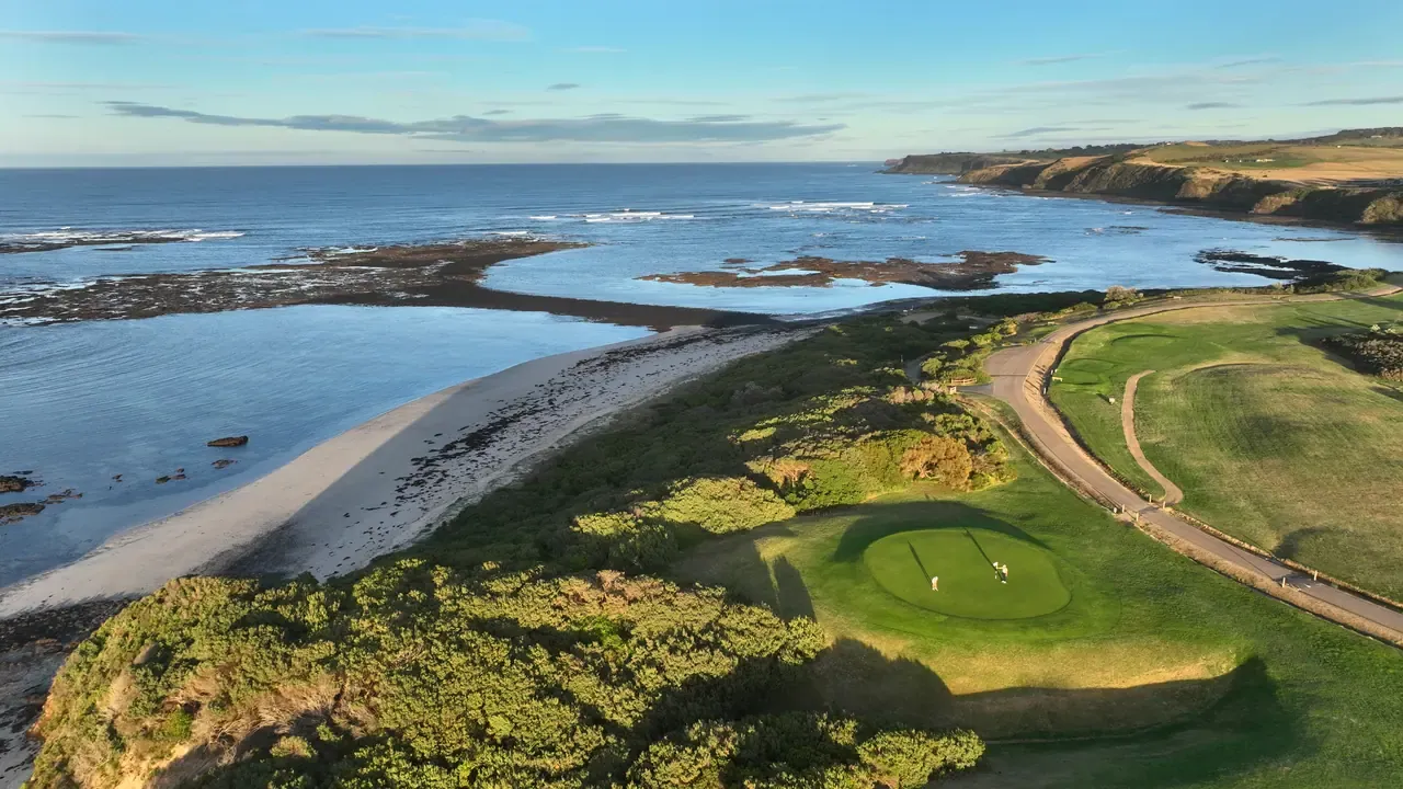 Aerial view of a headland golf green above a curved beach and tidal reef, with a cart path winding along the cliff edge.