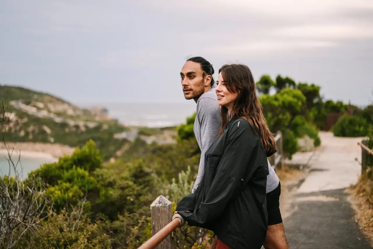 Two people lean on a timber rail on a coastal path at Point Nepean, looking out over scrub towards the water