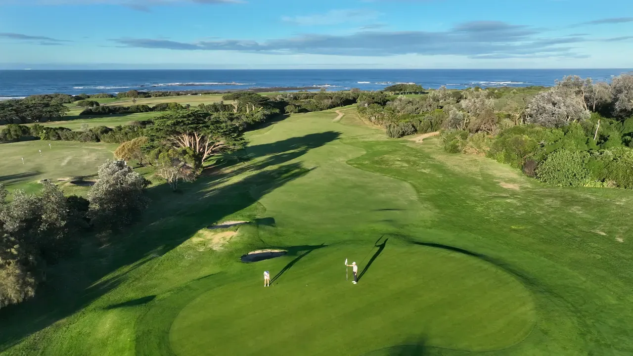 Aerial view down a fairway to a green at Flinders Golf Course, with two golfers, bunkers and the ocean on the horizon.