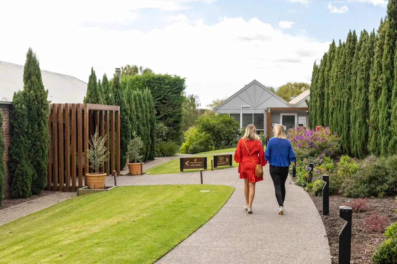Two visitors walk a path past lawn, cypress trees and flowering garden beds towards the gabled cellar door at Crittenden Estate