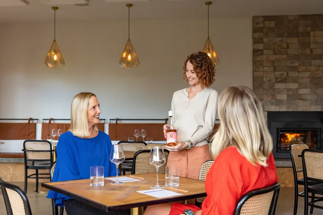 A host presents a bottle of rose to two seated guests in a tasting room with pendant lights and a stone fireplace
