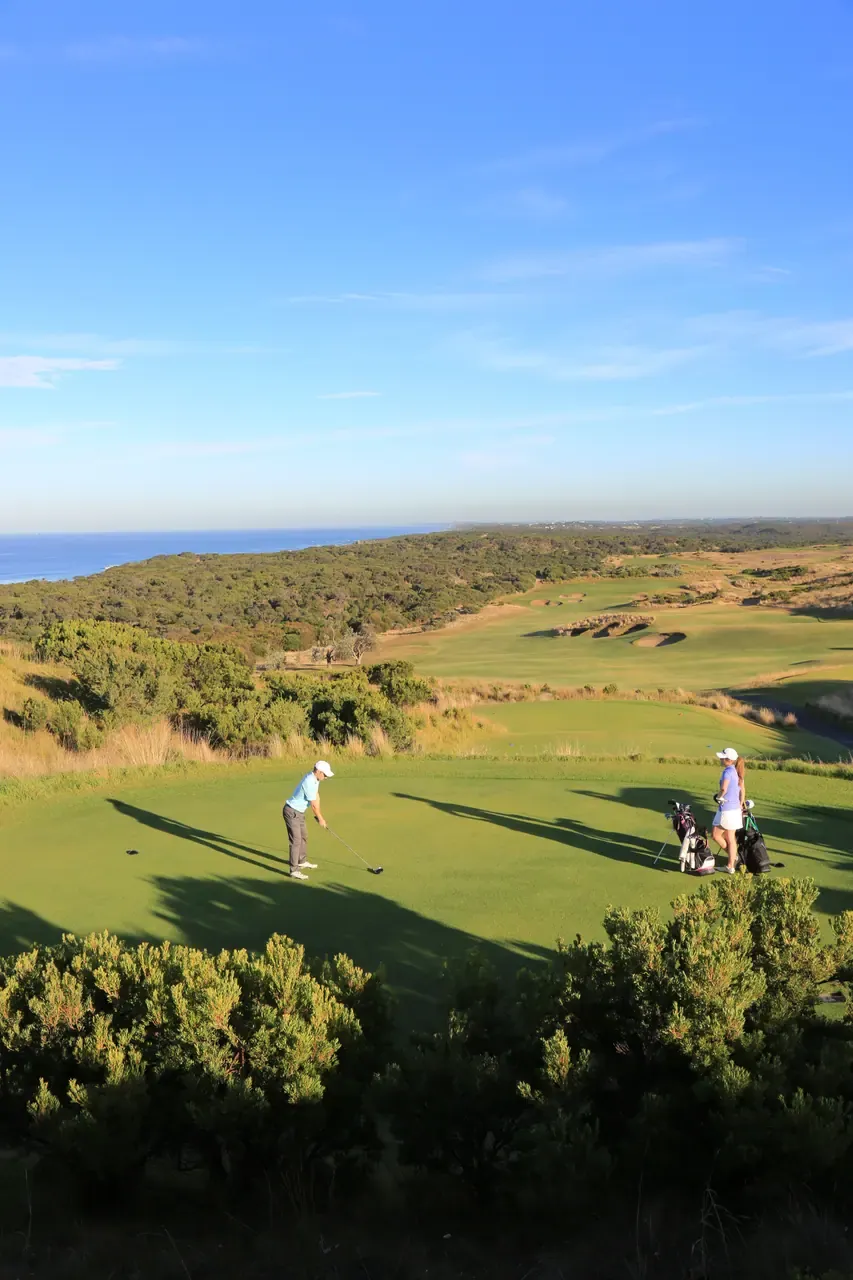 A golfer lines up a tee shot at The National Golf Club, a partner by the bags and fairways running toward the distant sea