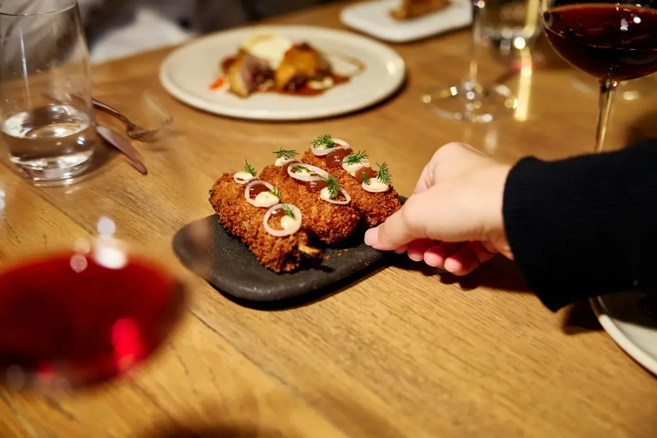 A hand reaches for crumbed croquettes topped with onion rings and dill on a slate board, with red wine, at Lindenderry