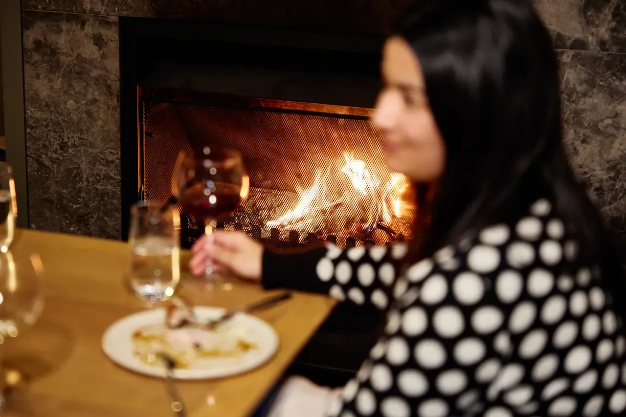 Glass of red wine and a dessert plate on a table in front of an open wood fire, a diner blurred in the foreground
