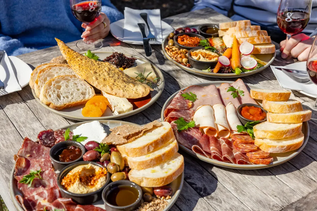 Shared platters of cured meats, bread, dips, olives and cheese on a sunlit outdoor table with red wine at Green Olive at Red Hill