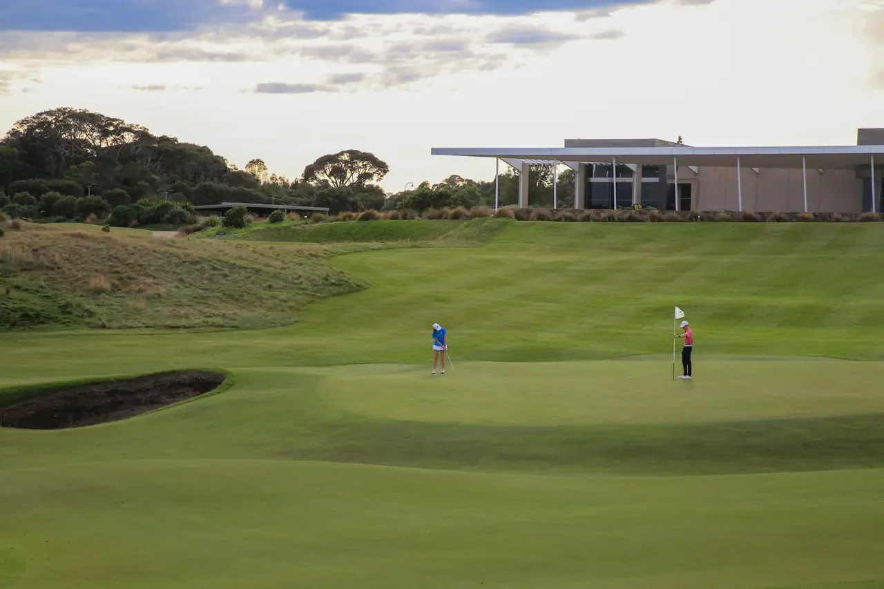 Two golfers on a green beside a bunker at Moonah Links Golf Course, Fingal, with the low modern clubhouse on the rise behind.