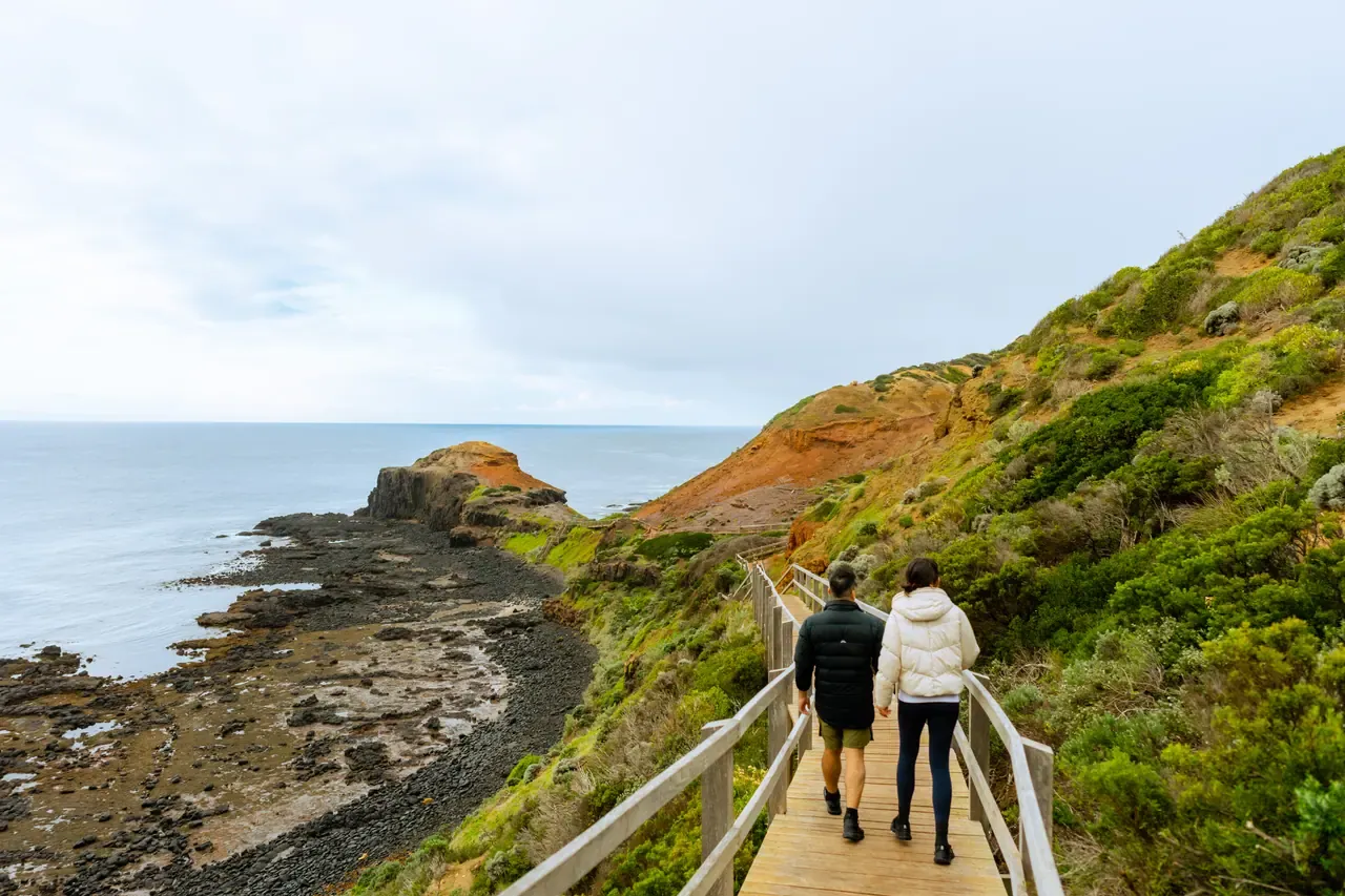 Two walkers head down the Cape Schanck Boardwalk towards a rocky basalt shore and flat-topped headland