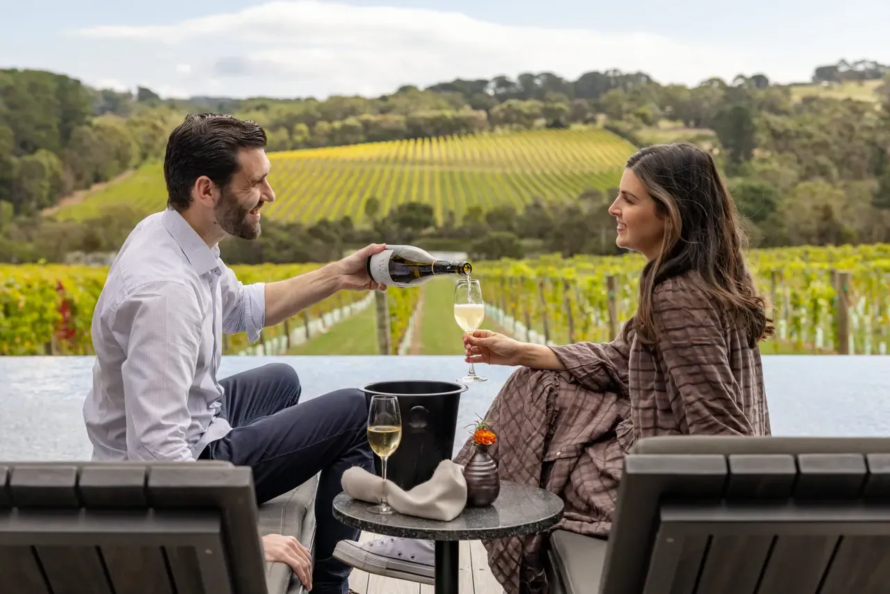 A man pours white wine for a woman on the terrace at Jackalope Hotel, with rows of vines and a hillside vineyard beyond