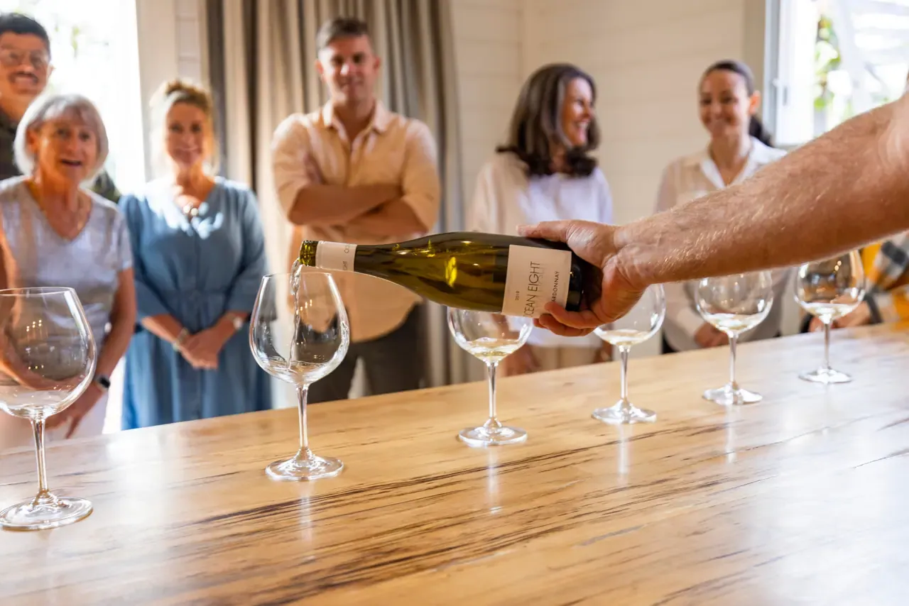 Chardonnay is poured into a row of glasses on a timber bench as a group waits at the Ocean Eight tasting room