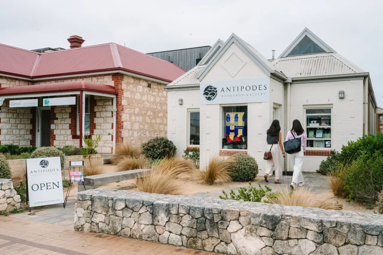 Two women walk towards the white facade of Antipodes Bookshop and Gallery in Sorrento, next to a limestone cottage and an Open sign