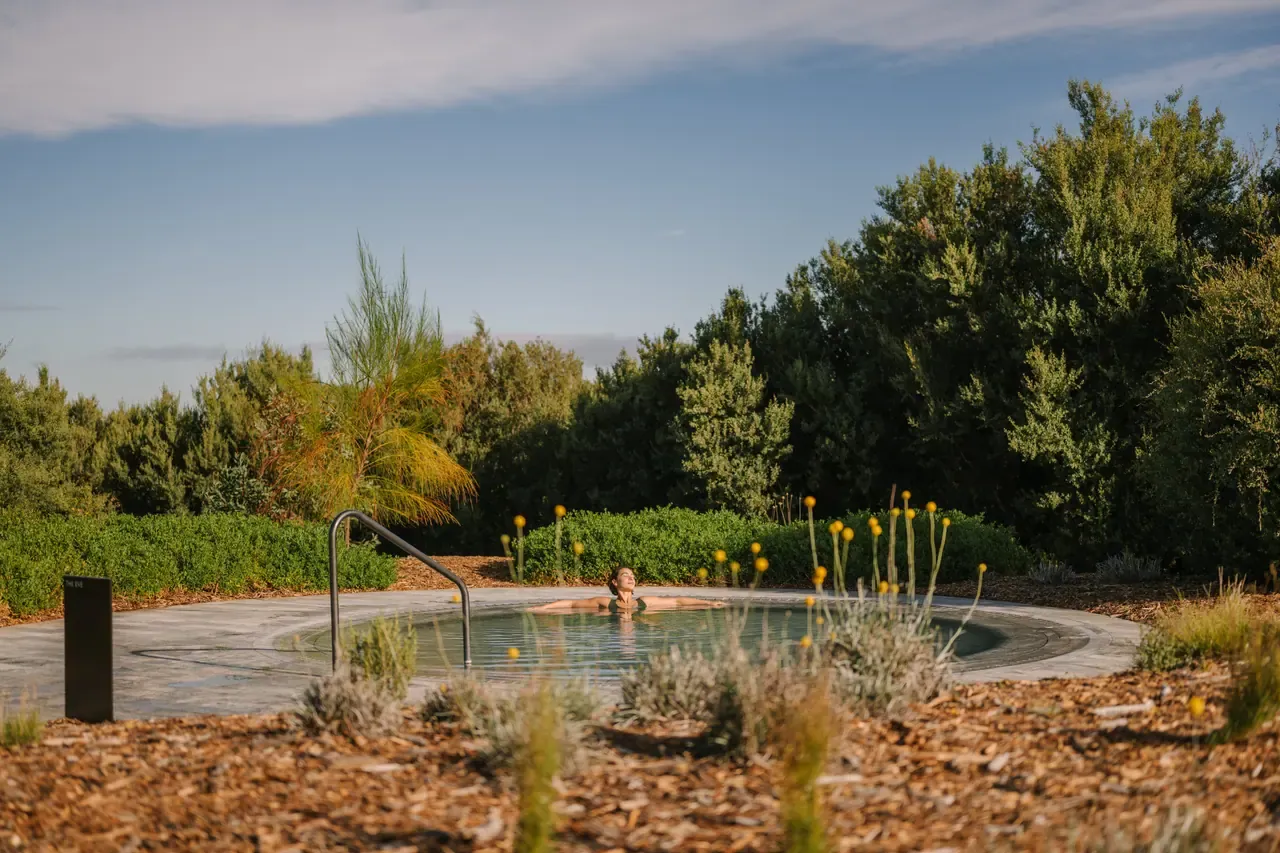 A single bather in a round outdoor hot pool framed by tall shrubs and yellow wildflowers at Alba Thermal Springs